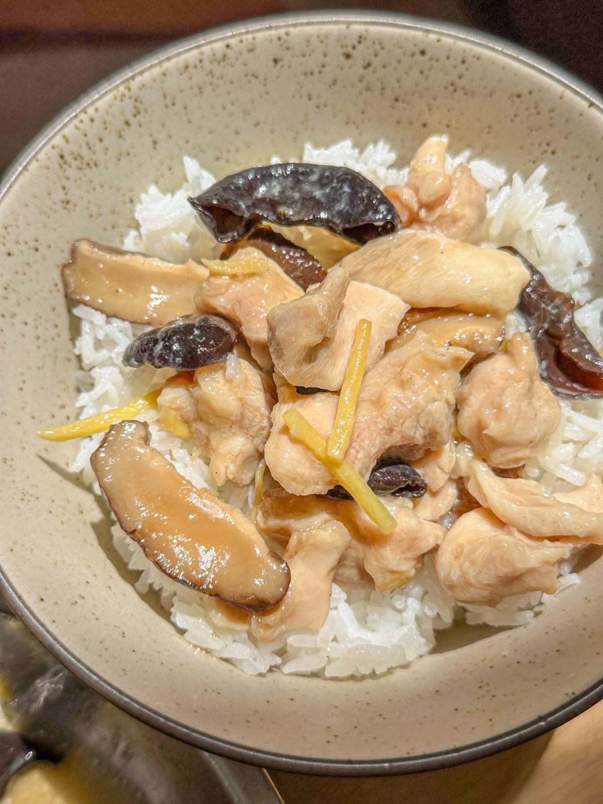 A bowl of white rice with steamed chicken, sliced shiitake mushrooms, wood ear mushrooms and ginger strips.