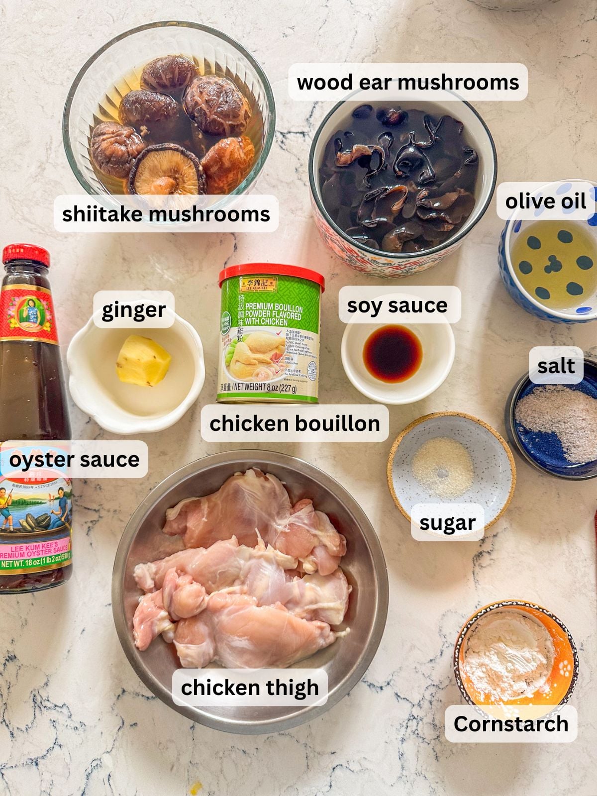 Ingredients for Chinese steamed chicken with mushrooms laid out on a white table countertop including shiitake mushrooms, wood ear mushrooms, ginger, chicken bouillon, chicken thigh, cornstarch, oyster sauce, soy sauce, salt and sugar.