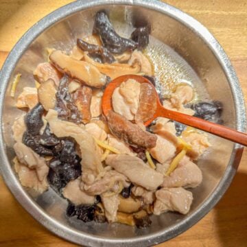 A bowl of steamed chicken with sliced shiitake mushrooms, wood ear mushrooms and ginger strips being scooped with a spoon.