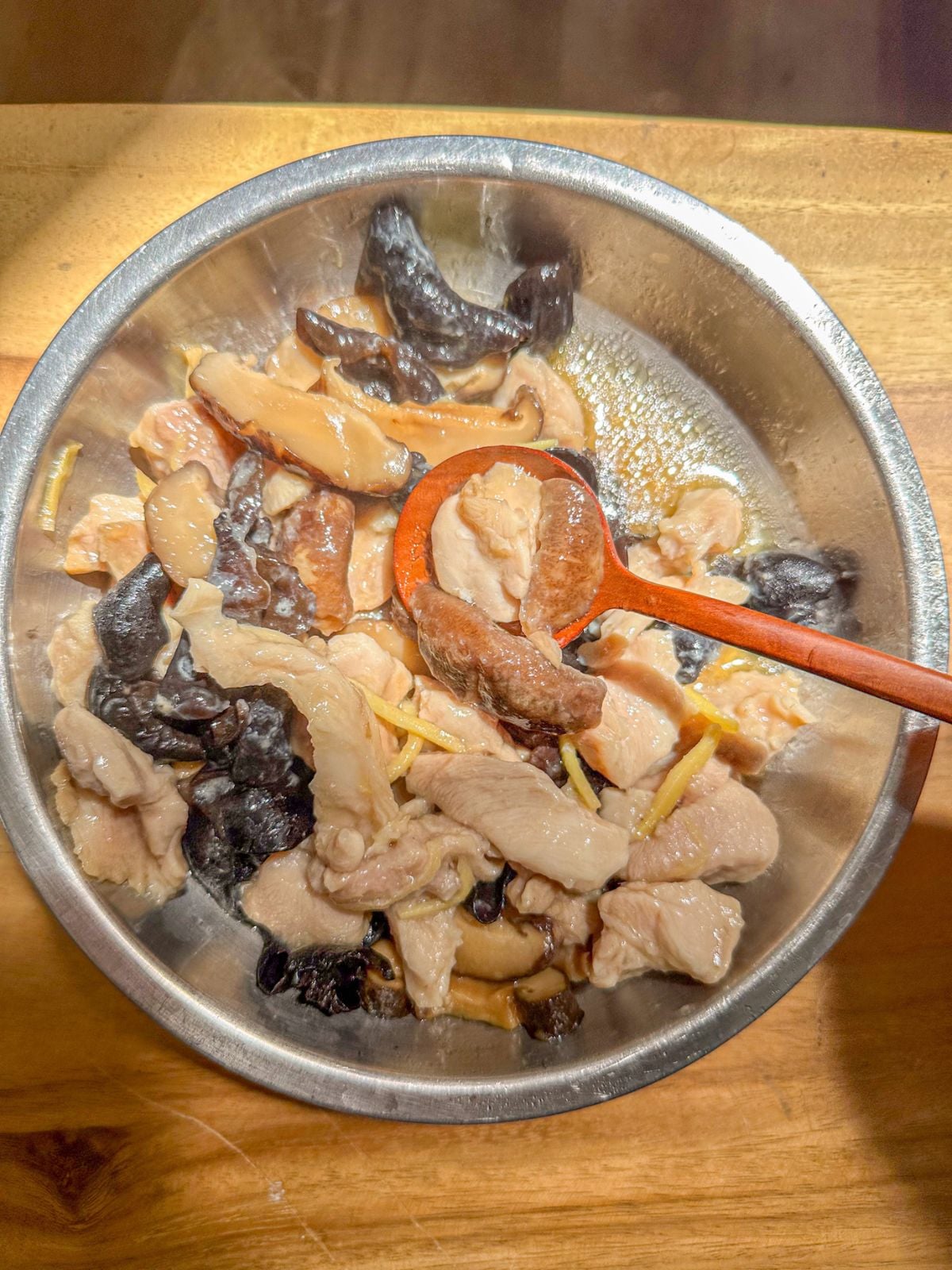 A bowl of steamed chicken with sliced shiitake mushrooms, wood ear mushrooms and ginger strips being scooped with a spoon.