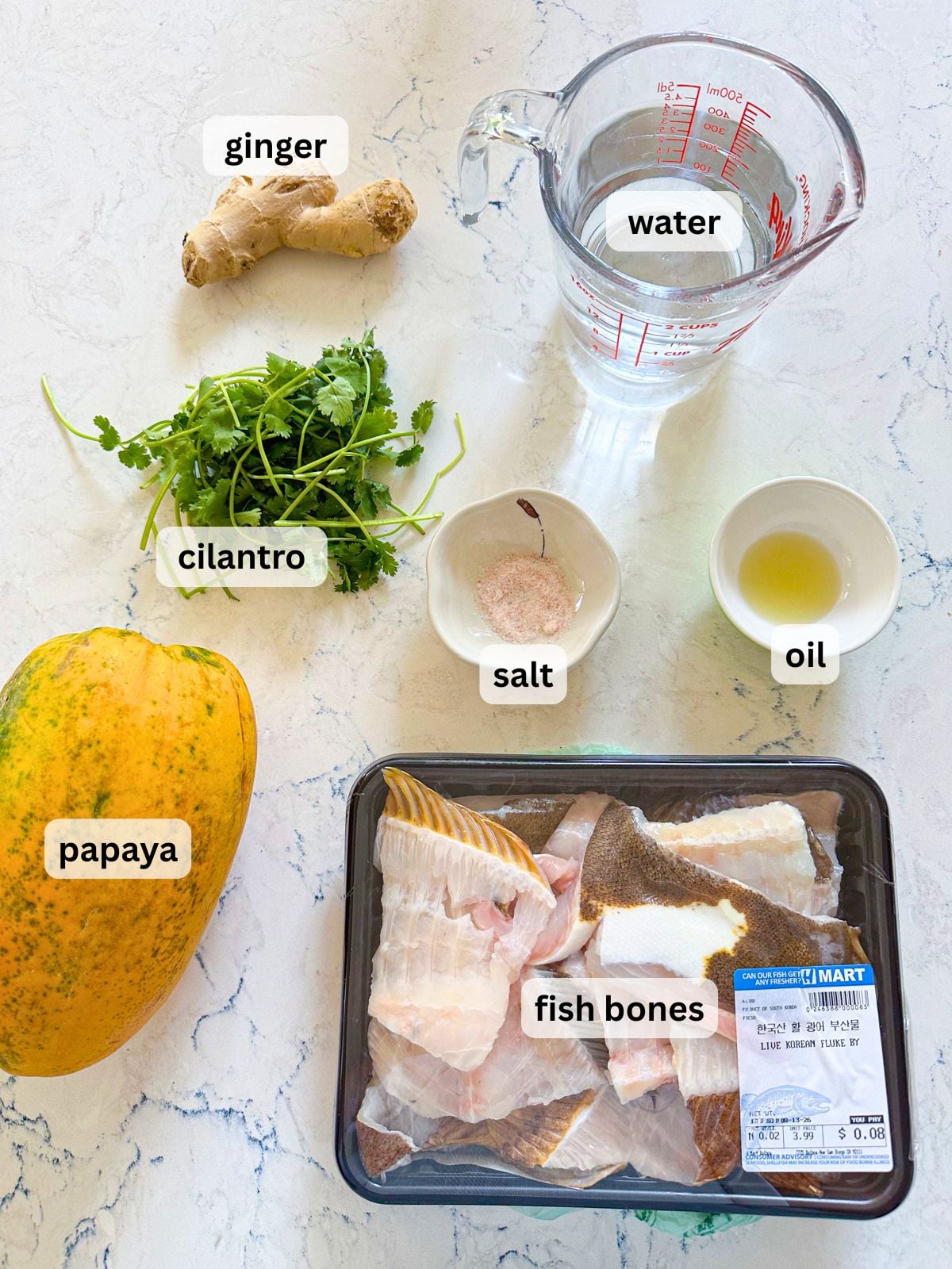 Fish and papaya soup ingredients laid out on a table countertop including ginger, cilantro, water, salt, oil, papaya and fish bones.