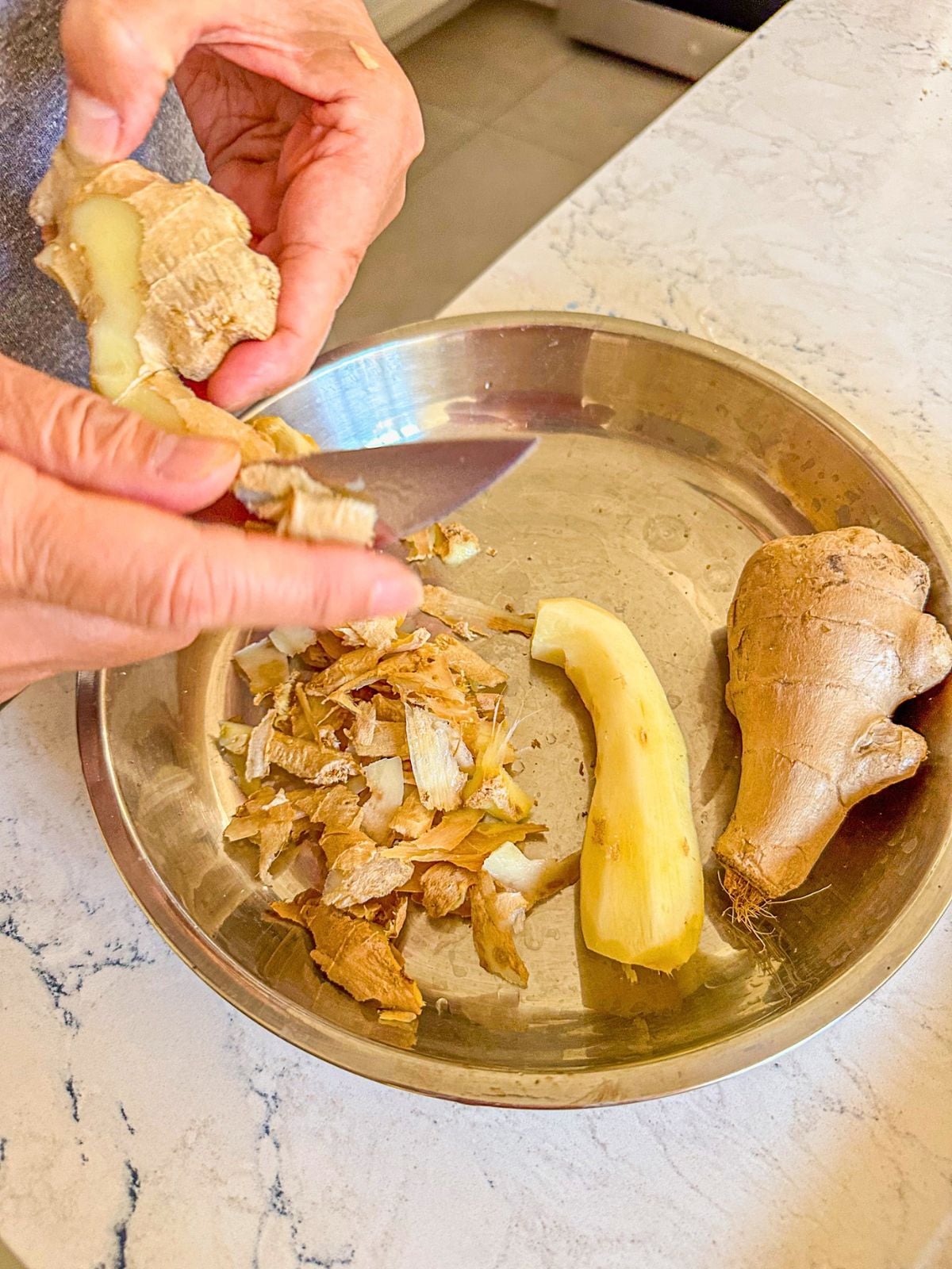 Holding half-peeled ginger beside a bowl of peeled and unpeeled ginger, with ginger peelings on the side.