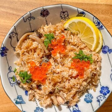 A plate of salmon and mushroom rice topped with green onions and fish roe, served with lemon slices on the side.