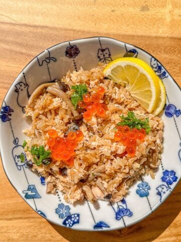 A plate of salmon and mushroom rice topped with green onions and fish roe, served with lemon slices on the side.