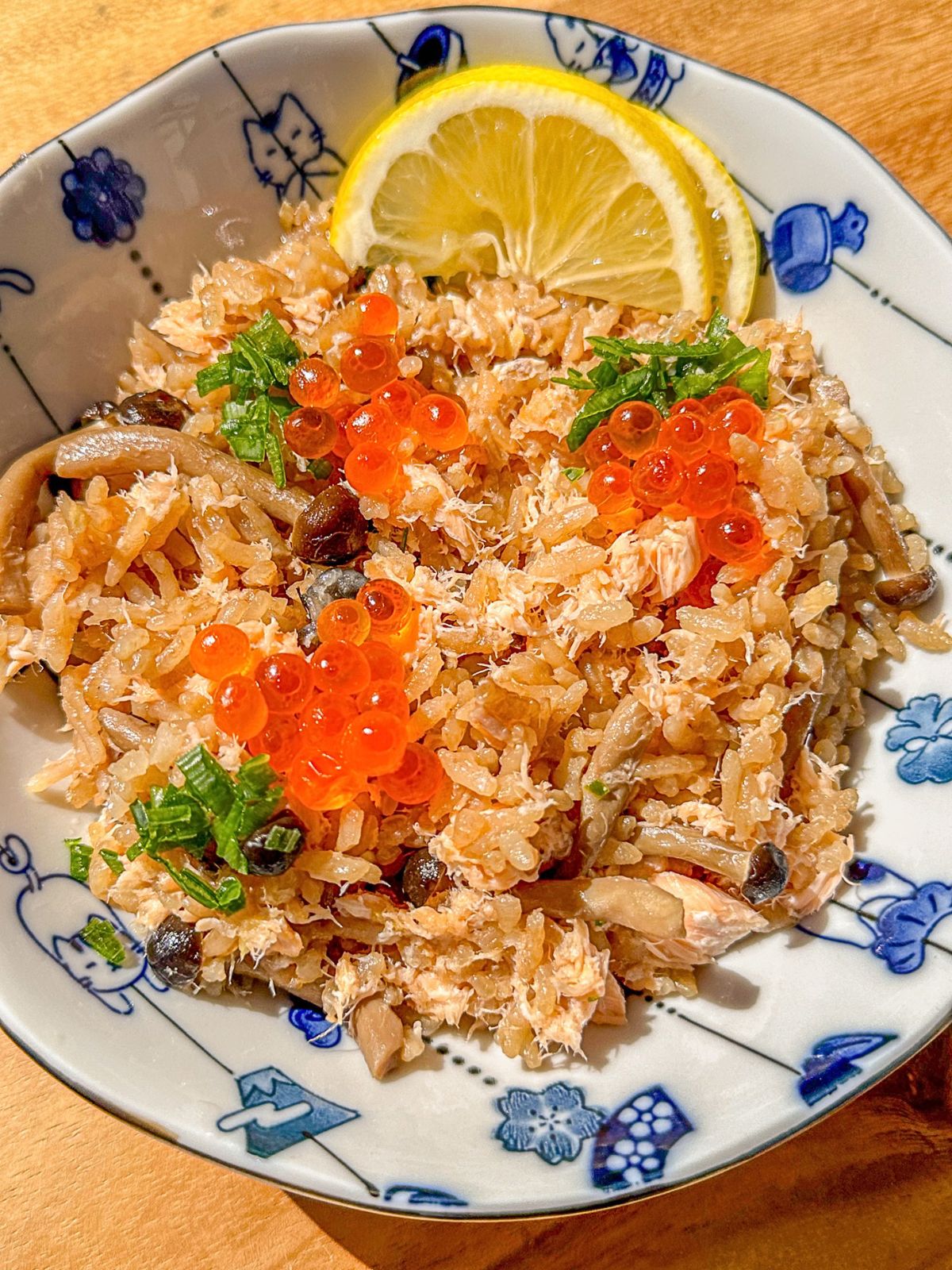 A plate of salmon and mushroom rice topped with green onions and fish roe, served with lemon slices on the side.