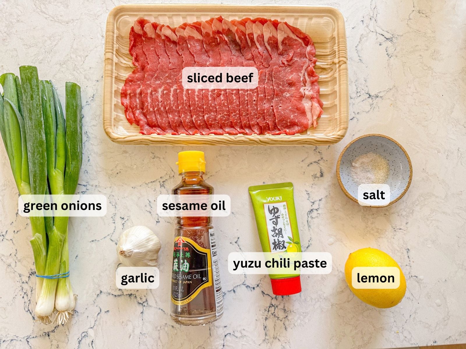 Green onions, sliced beef, garlic, lemon, sesame oil and yuzu kosho on a counter.