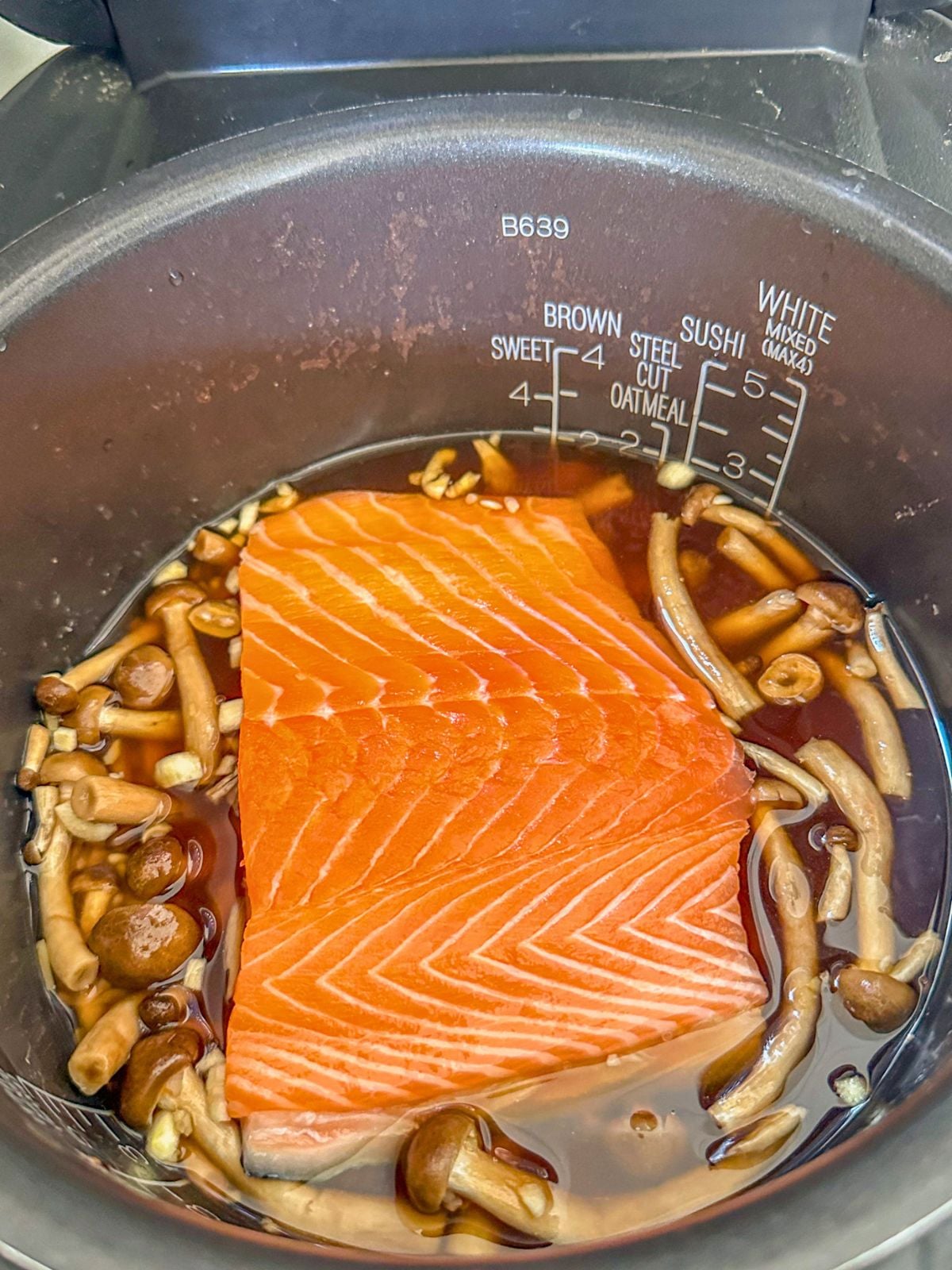 A pot of water with mushrooms, ginger, minced garlic, shallots, dark soy sauce, soy sauce, and oyster sauce, topped with salmon in the center.